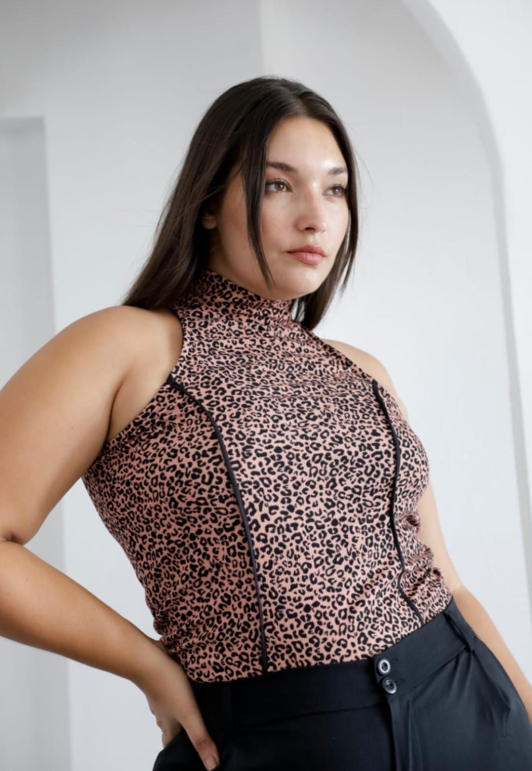 Mujer con un top con estampado de leopardo y pantalones negros, con la mano en el bolsillo, delante de una pared blanca.