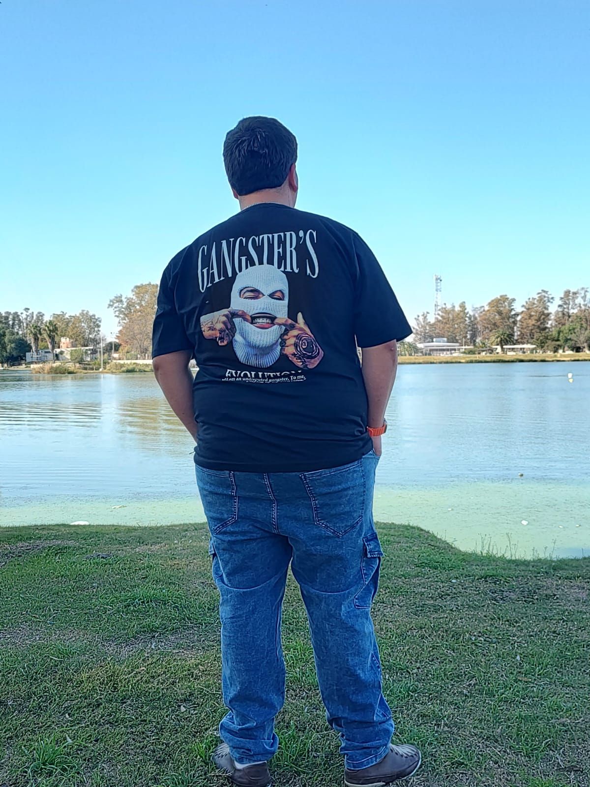 Hombre de pie sobre el césped junto a un lago, con camisa negra, vaqueros y zapatillas deportivas, sonriendo. Día soleado.