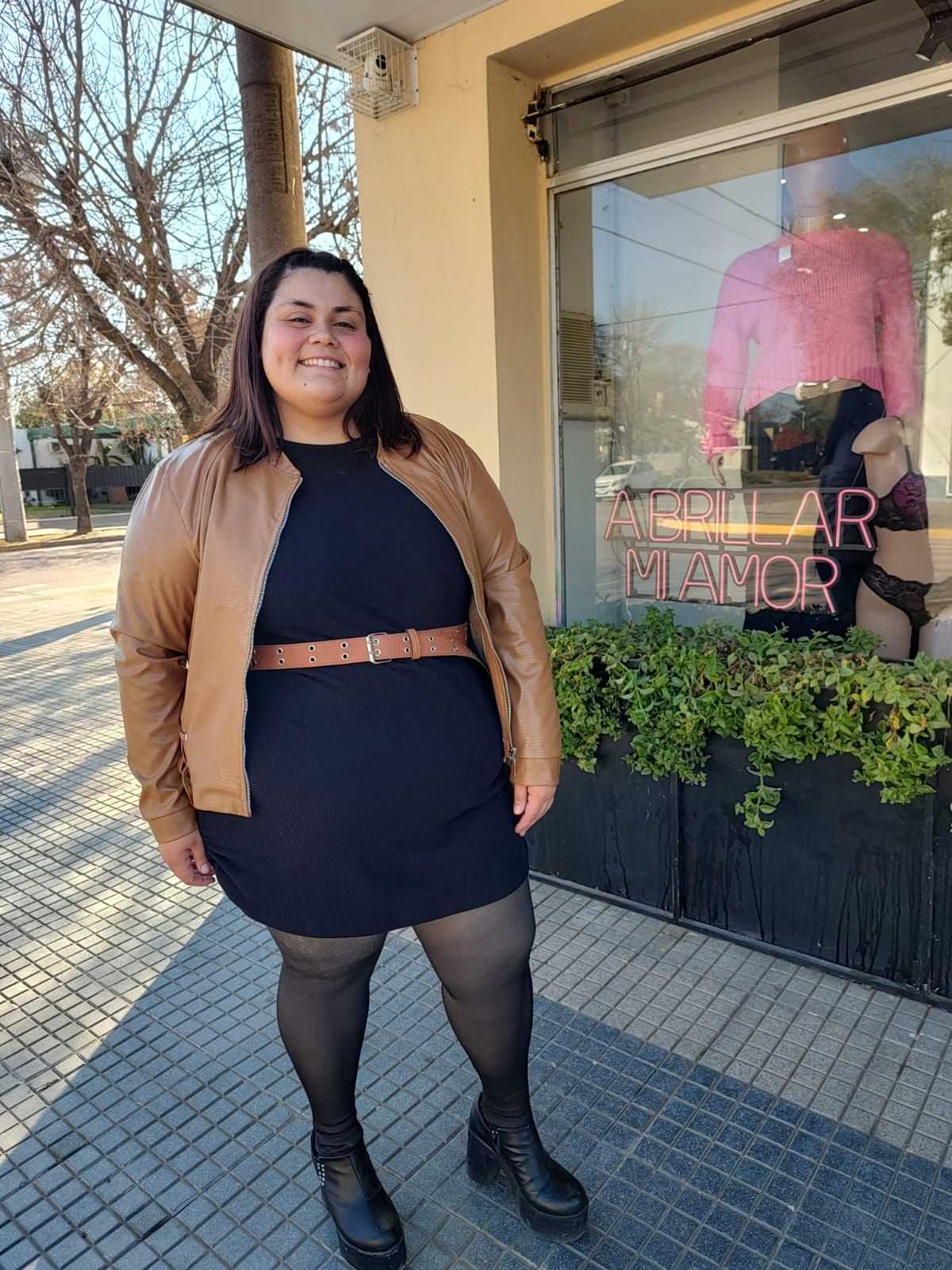 Una mujer con vestido negro y chaqueta marrón sonríe delante de una tienda.