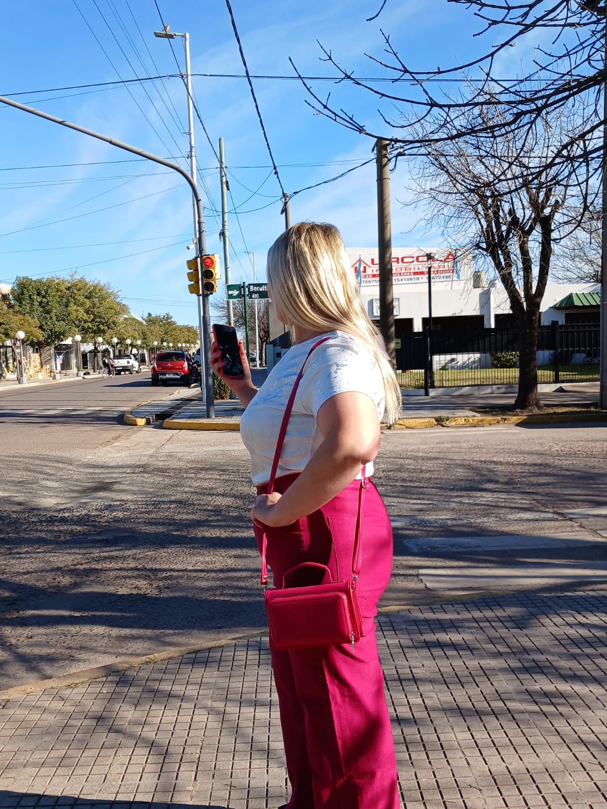 Mujer con camiseta roja y pantalones negros de pie en una acera, mirando a la izquierda. Día soleado, fondo urbano.