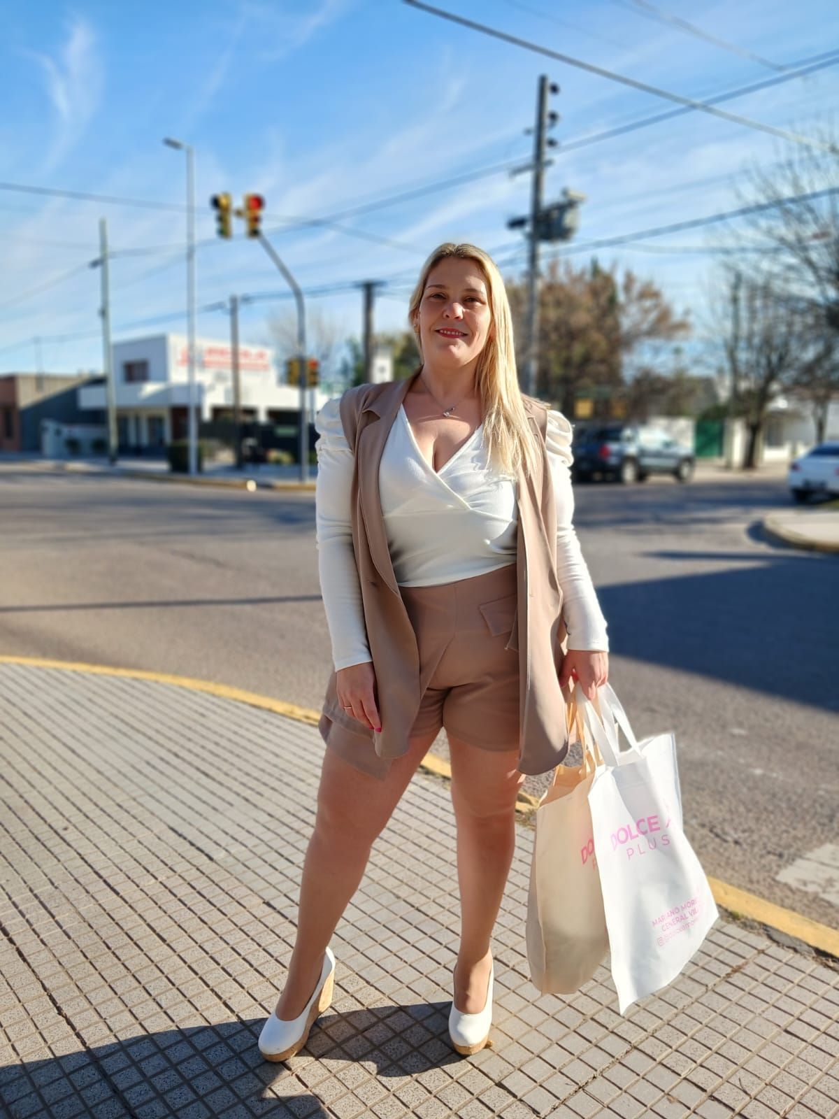 Mujer con traje corto beige sosteniendo bolsas, parada en una calle de la ciudad cerca de un cruce de peatones.