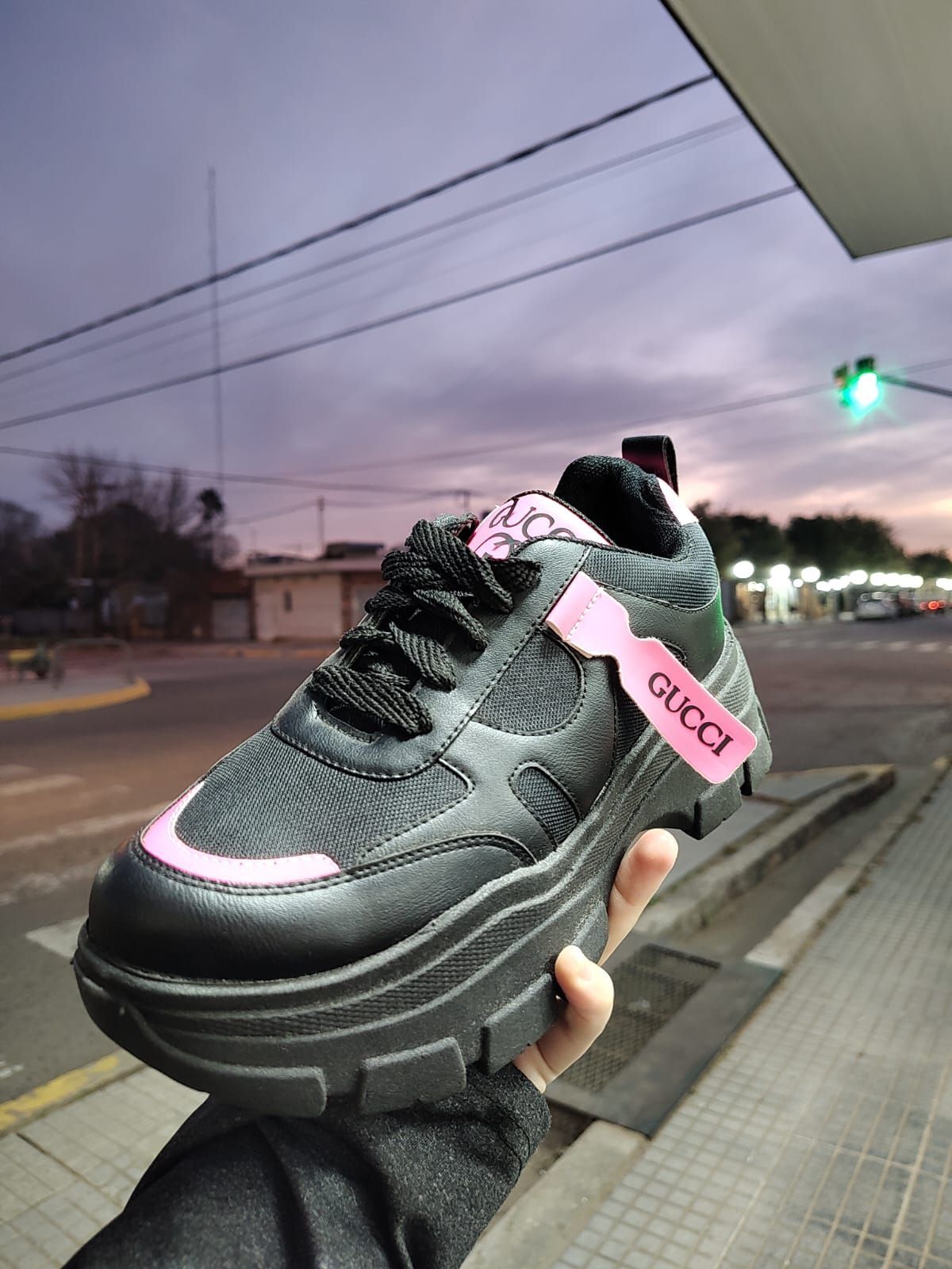 Persona sosteniendo una zapatilla Gucci blanca y rosa al aire libre, entorno nocturno.