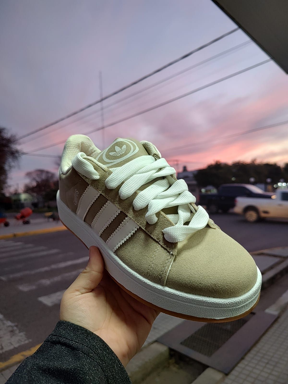 Mano sosteniendo una zapatilla Adidas negra con tres rayas, frente a una escena callejera al atardecer con un cielo oscuro.