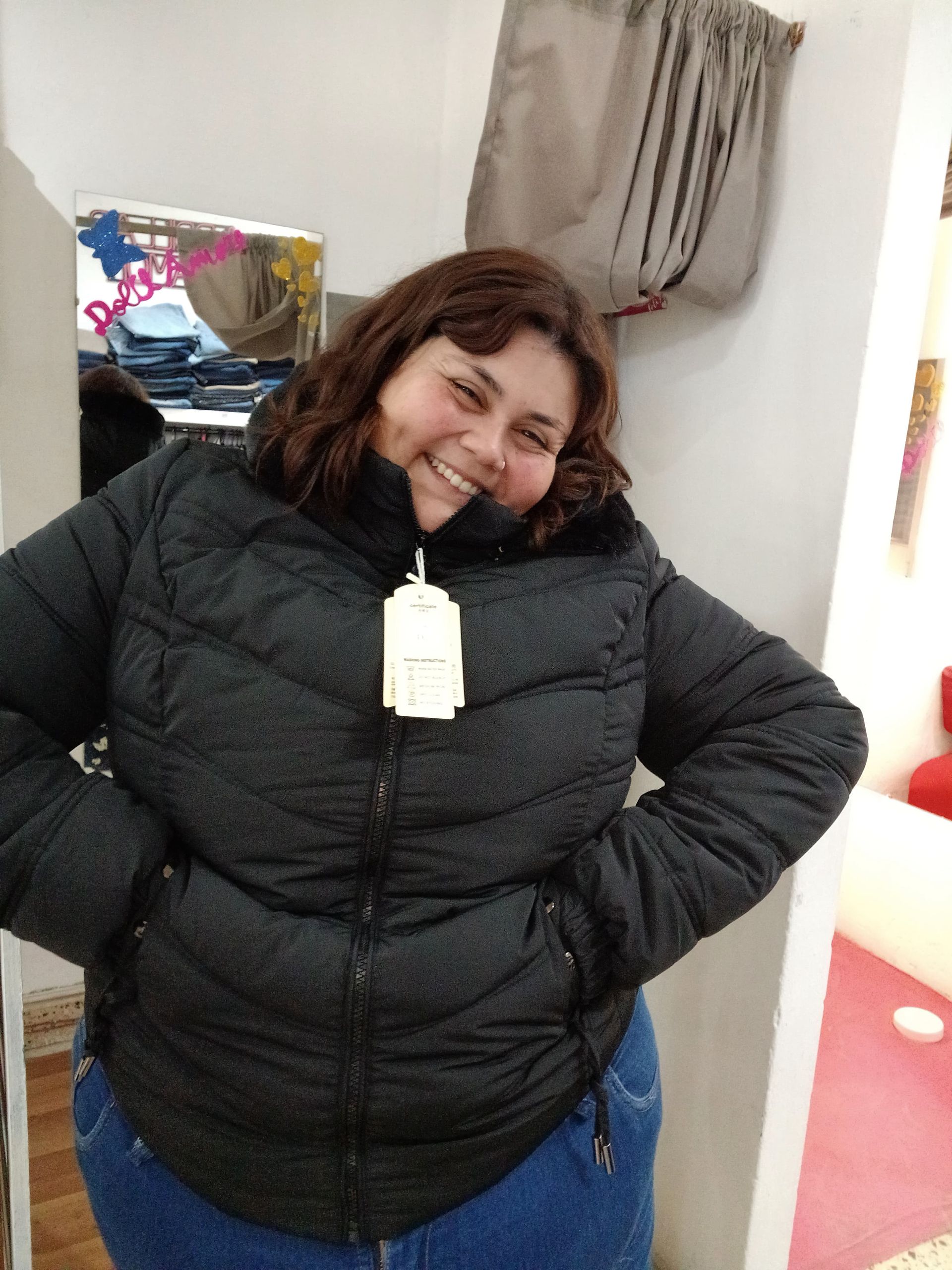 Mujer con una chaqueta acolchada de color marrón, sonriendo, de pie en una tienda de ropa, con las manos en los bolsillos.