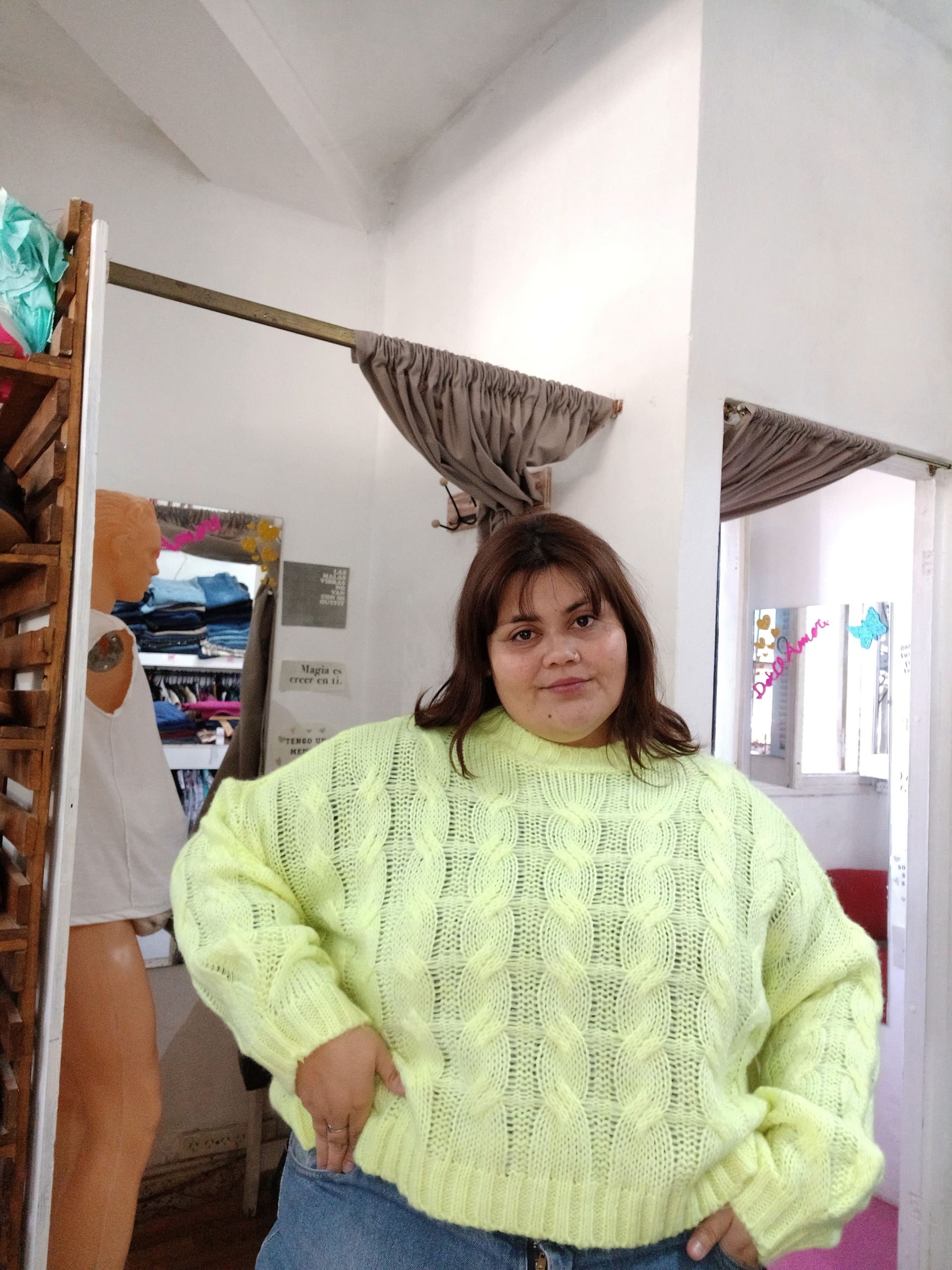 Una mujer con un suéter rosa brillante levanta la vista y se toca el pelo. De pie cerca de una puerta, en el interior.