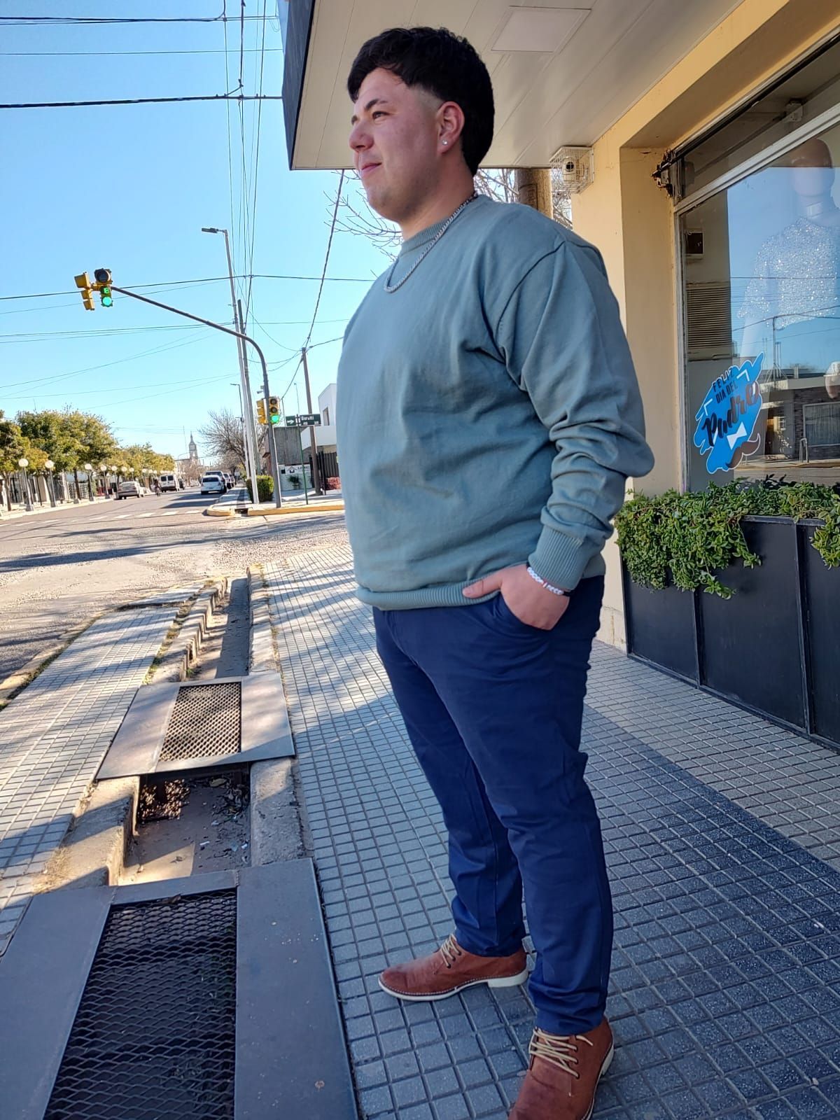Hombre con suéter azul verdoso y pantalones azul marino sonriendo, con las manos en los bolsillos