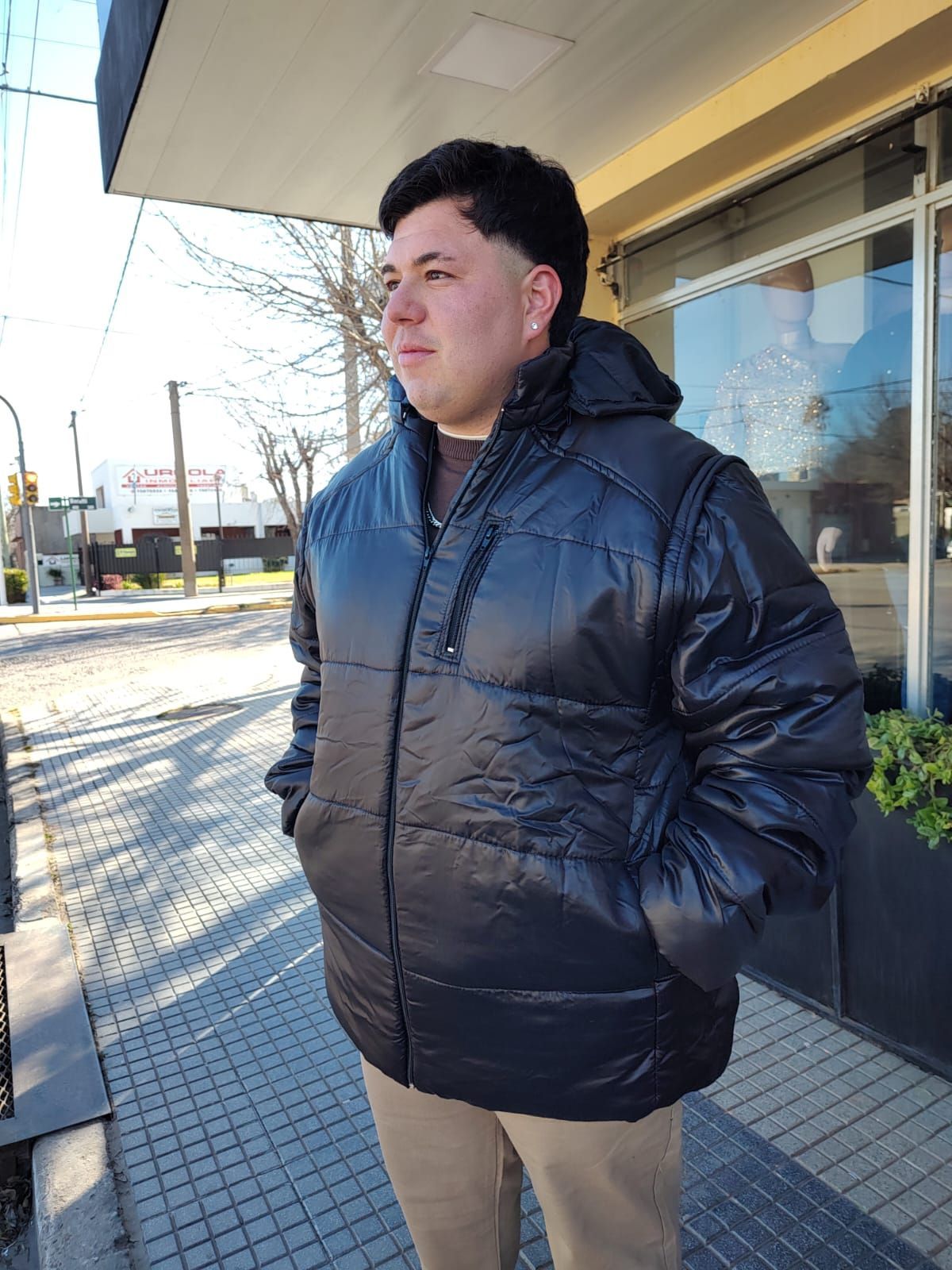 Hombre con una chaqueta negra inflada con capucha, de pie afuera de un edificio, con las manos en los bolsillos.