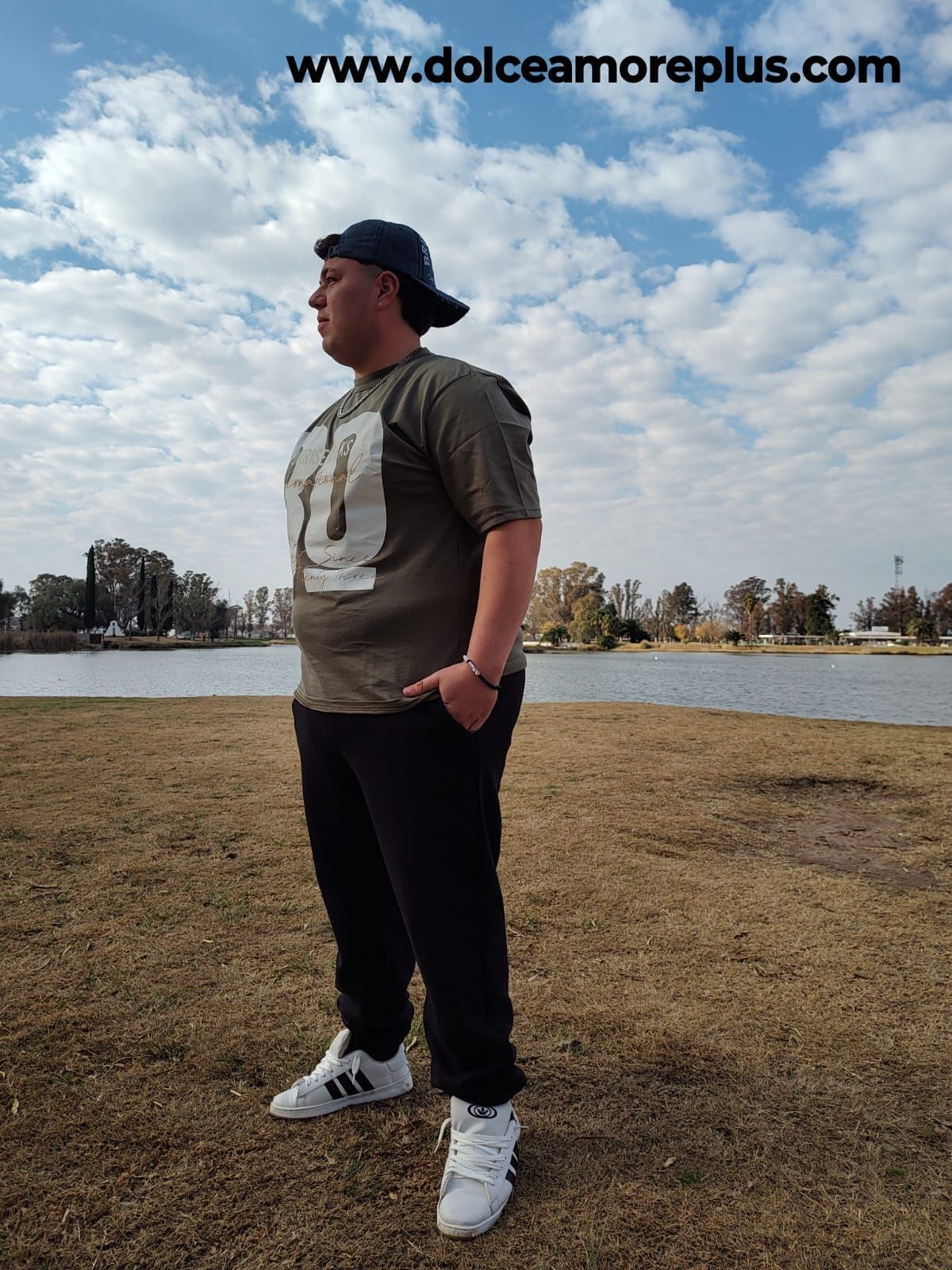 Dos personas con sombreros y ropa casual posan al aire libre, sonrientes, con un lago y un cielo nublado de fondo.