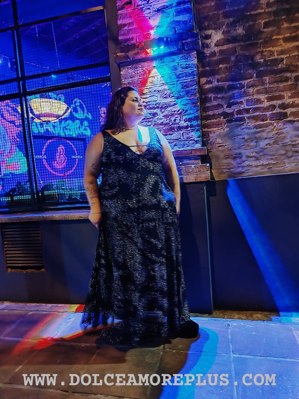 Una mujer con un vestido negro brillante está parada frente a una ventana, iluminada por una luz azul y violeta.