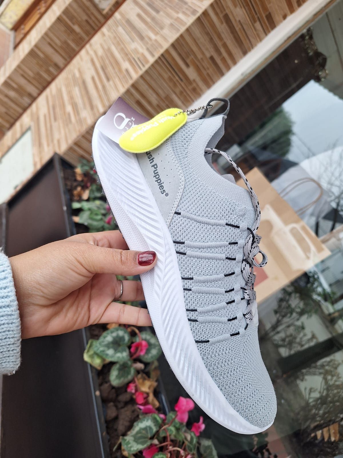 Zapatilla Hush Puppies gris en exposición, sostenida por una persona. Suela blanca, etiqueta amarilla.