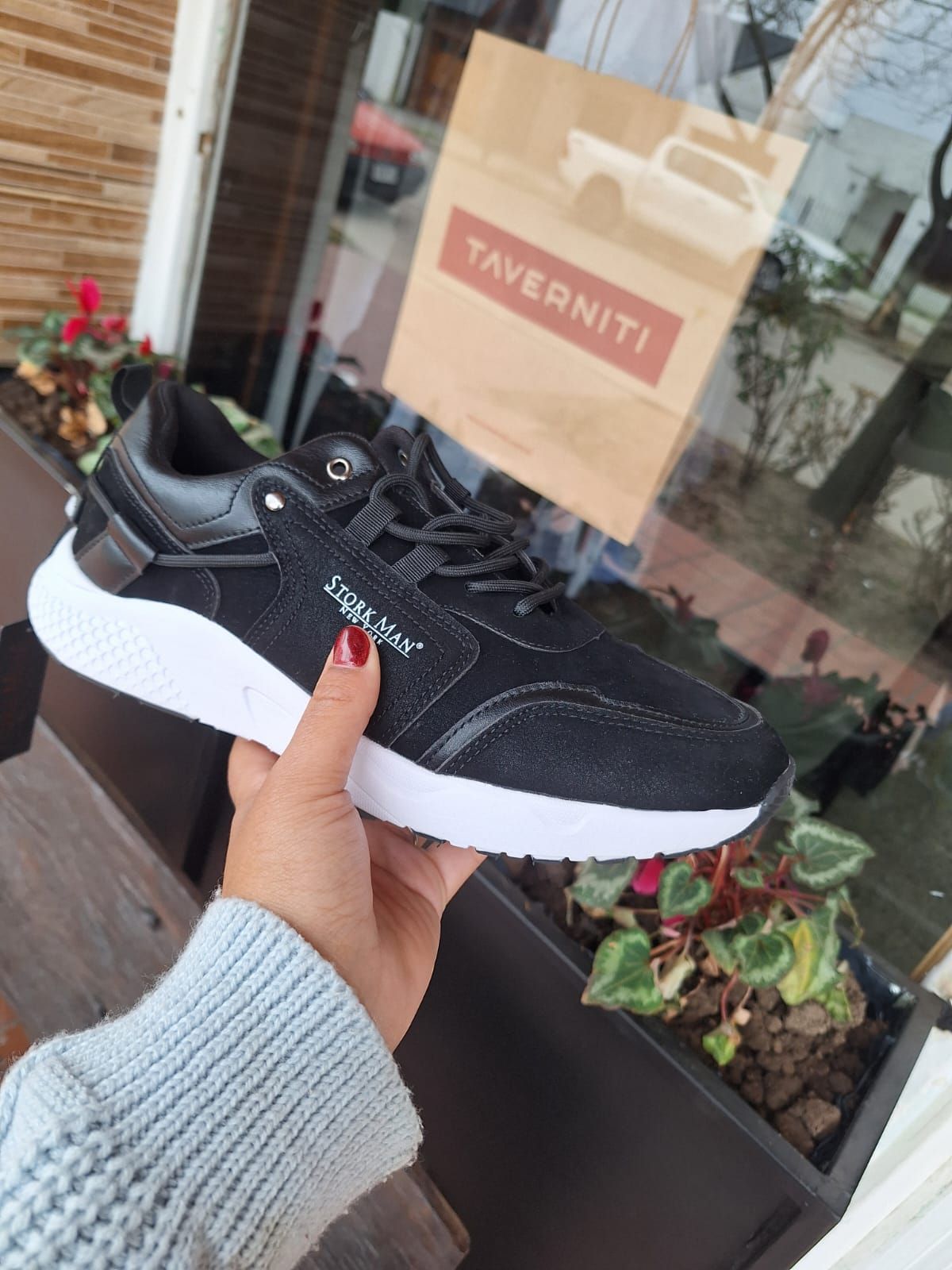 Zapatilla blanca y negra sostenida en la mano, frente a una ventana con una bolsa de papel y plantas.