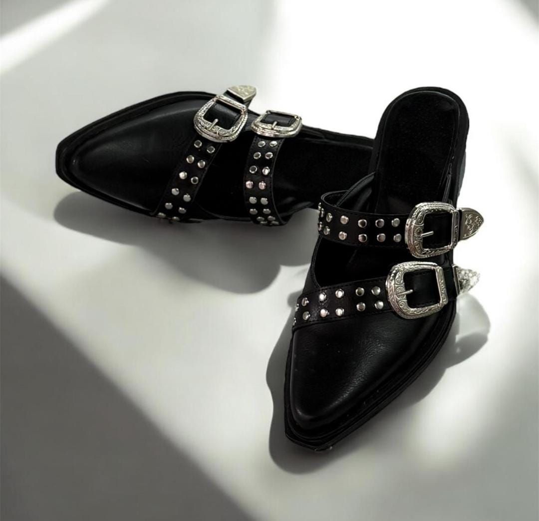 Zuecos de piel de color negro con hebillas y tachuelas plateadas, apoyados sobre una superficie reflectante.
