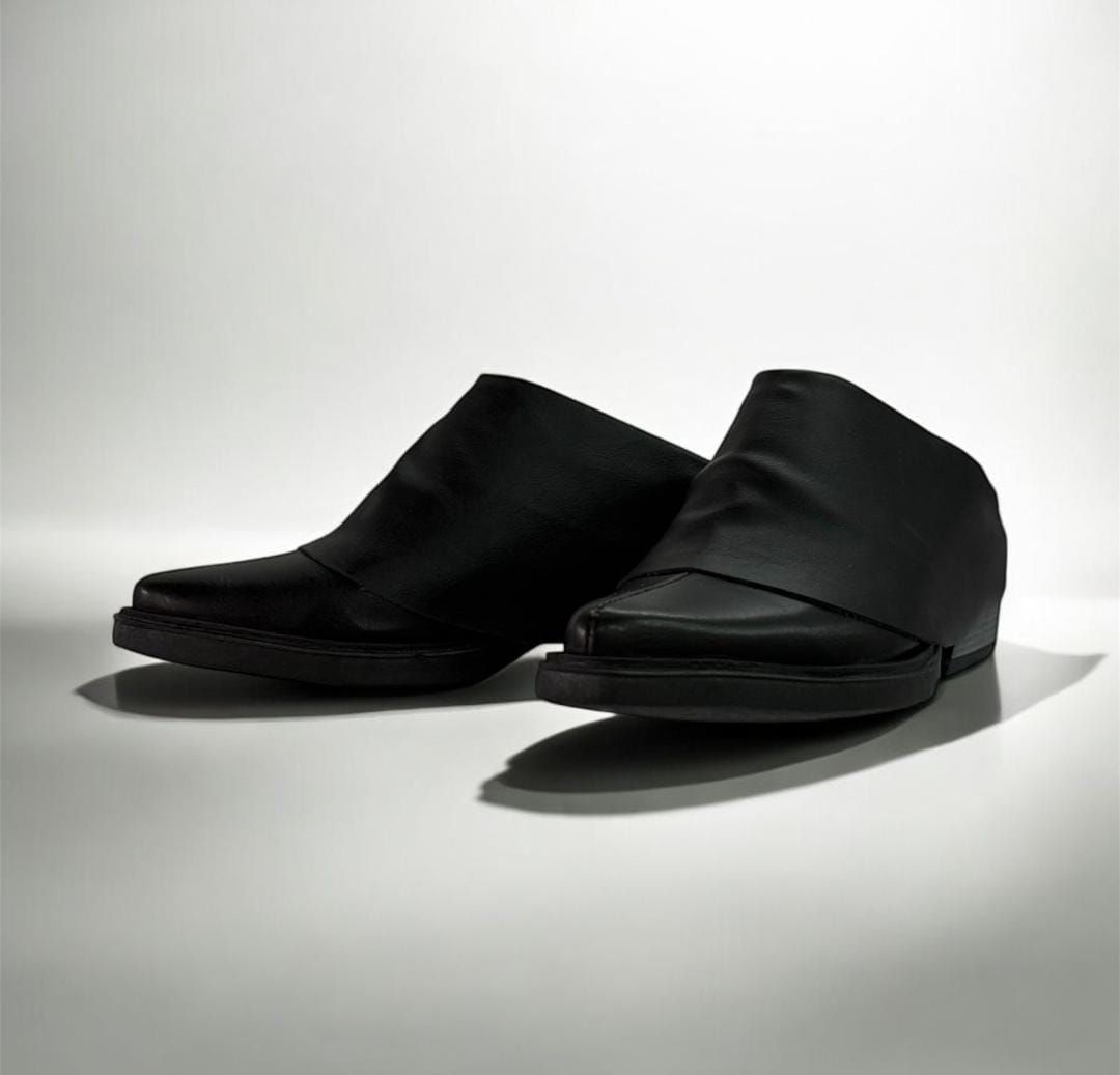 Zapatos sin cordones de piel negra sobre superficie reflectante blanca.