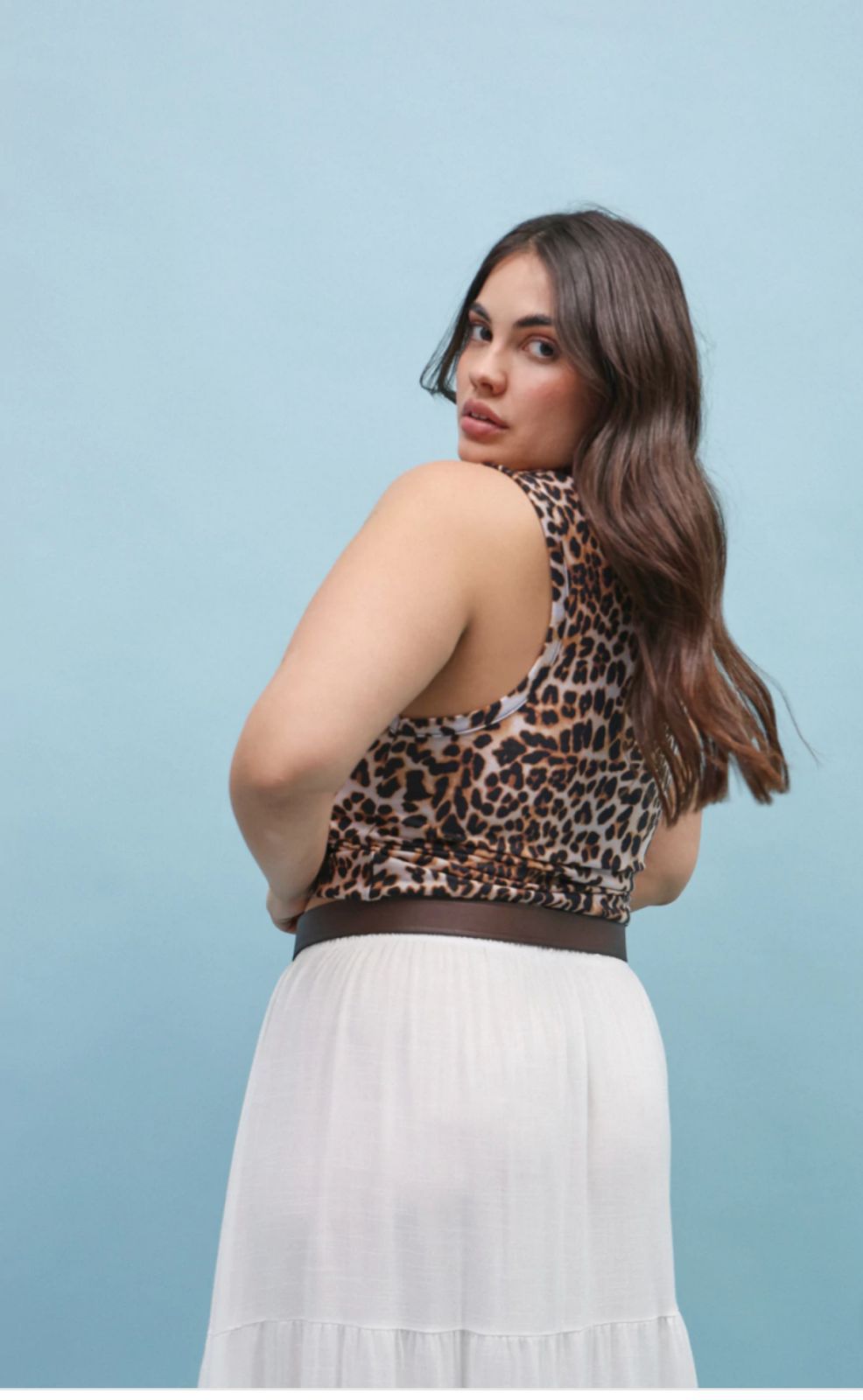Mujer con top corto con estampado de leopardo, falda blanca y cinturón marrón, parada frente a un fondo azul.
