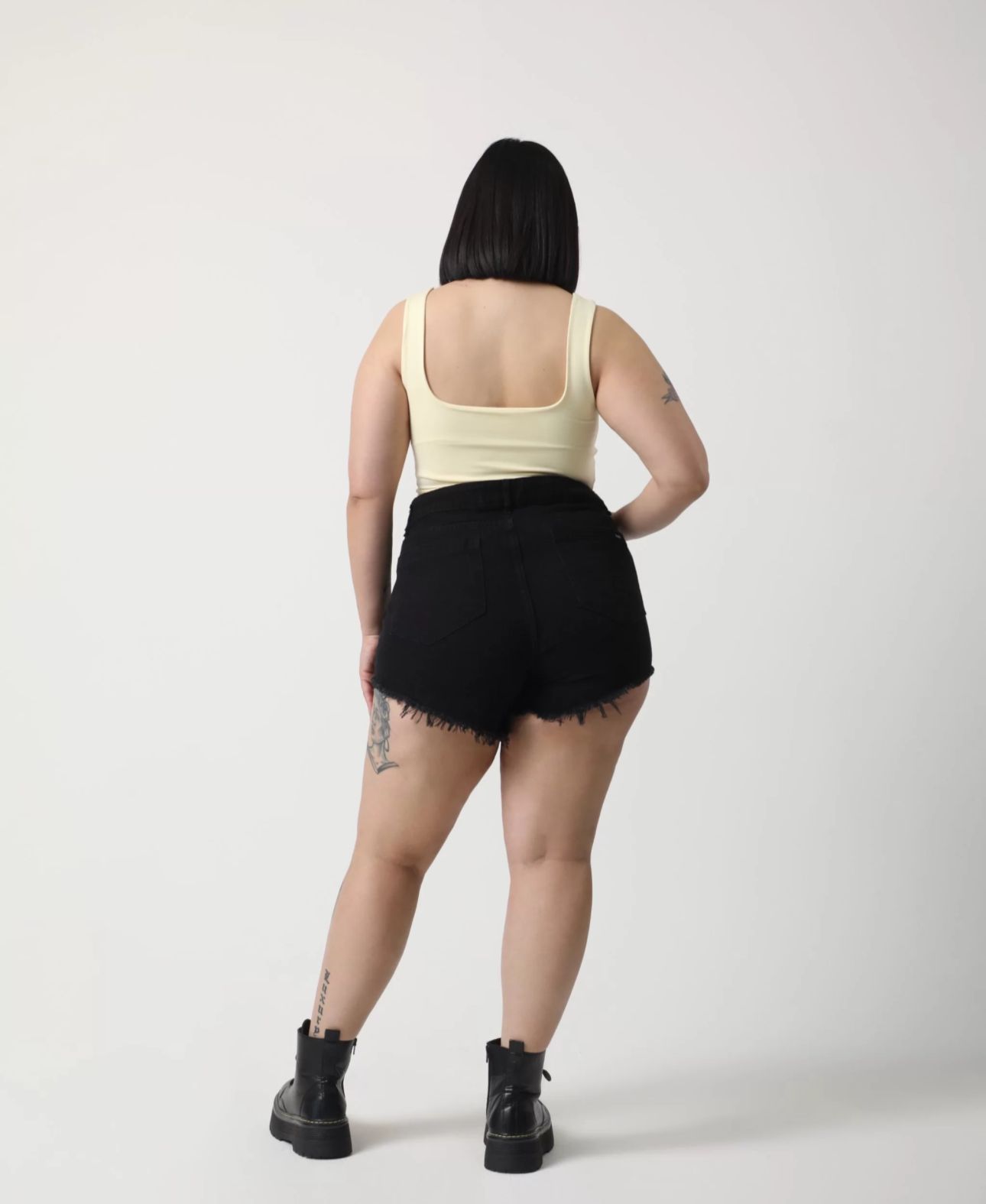 Mujer con una camiseta amarilla y pantalones cortos negros con tatuajes, de pie frente a un fondo blanco.