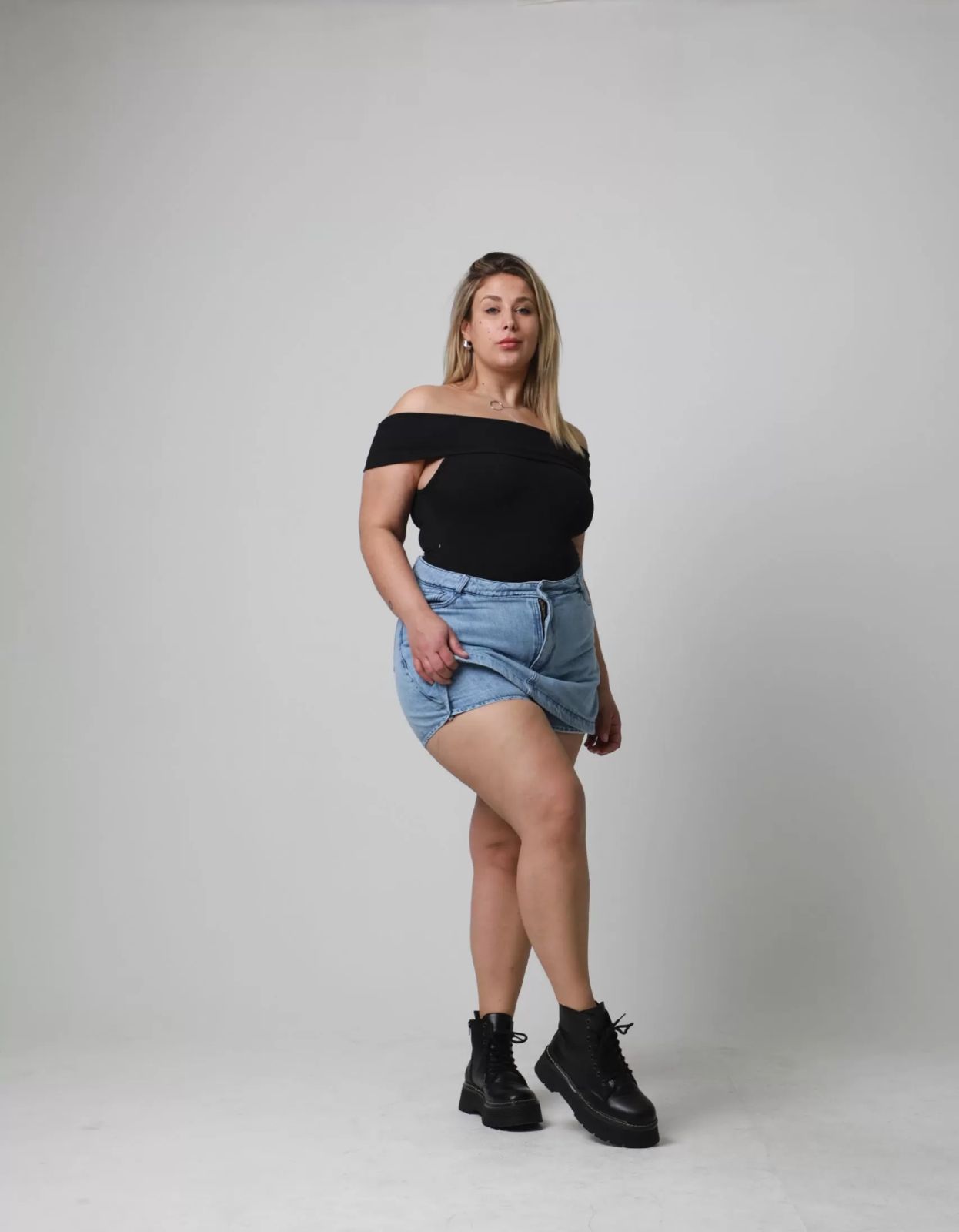 Mujer con top negro con hombros descubiertos, falda vaquera y botas negras posando en un estudio.
