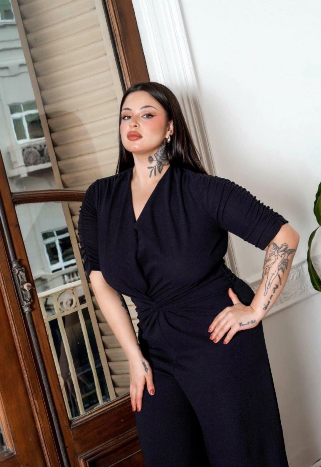 Mujer con un mono negro, de pie cerca de una puerta y un sofá, con los brazos a los costados.