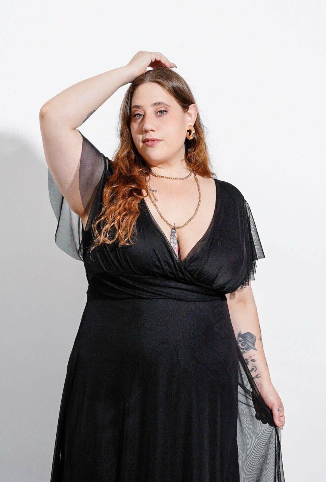 Mujer con un vestido negro con mangas transparentes, tacones negros, sosteniendo su vestido, de pie frente a un fondo blanco.