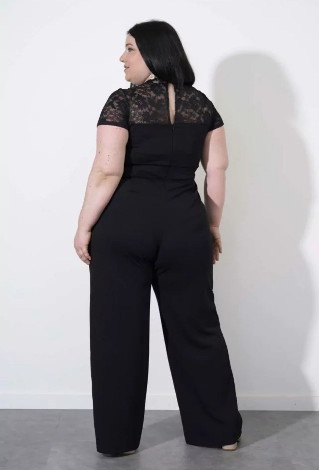 Mujer con un mono negro apoyada contra una pared blanca, mirando hacia un lado.