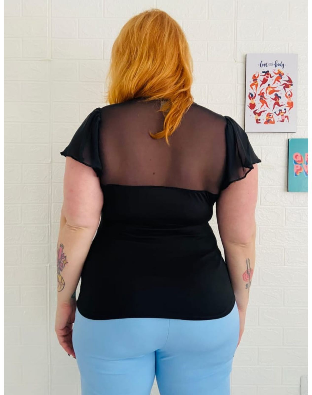 Mujer con top negro y pantalones azules, sujetándose el cabello, parada frente a una pared de ladrillos blancos.
