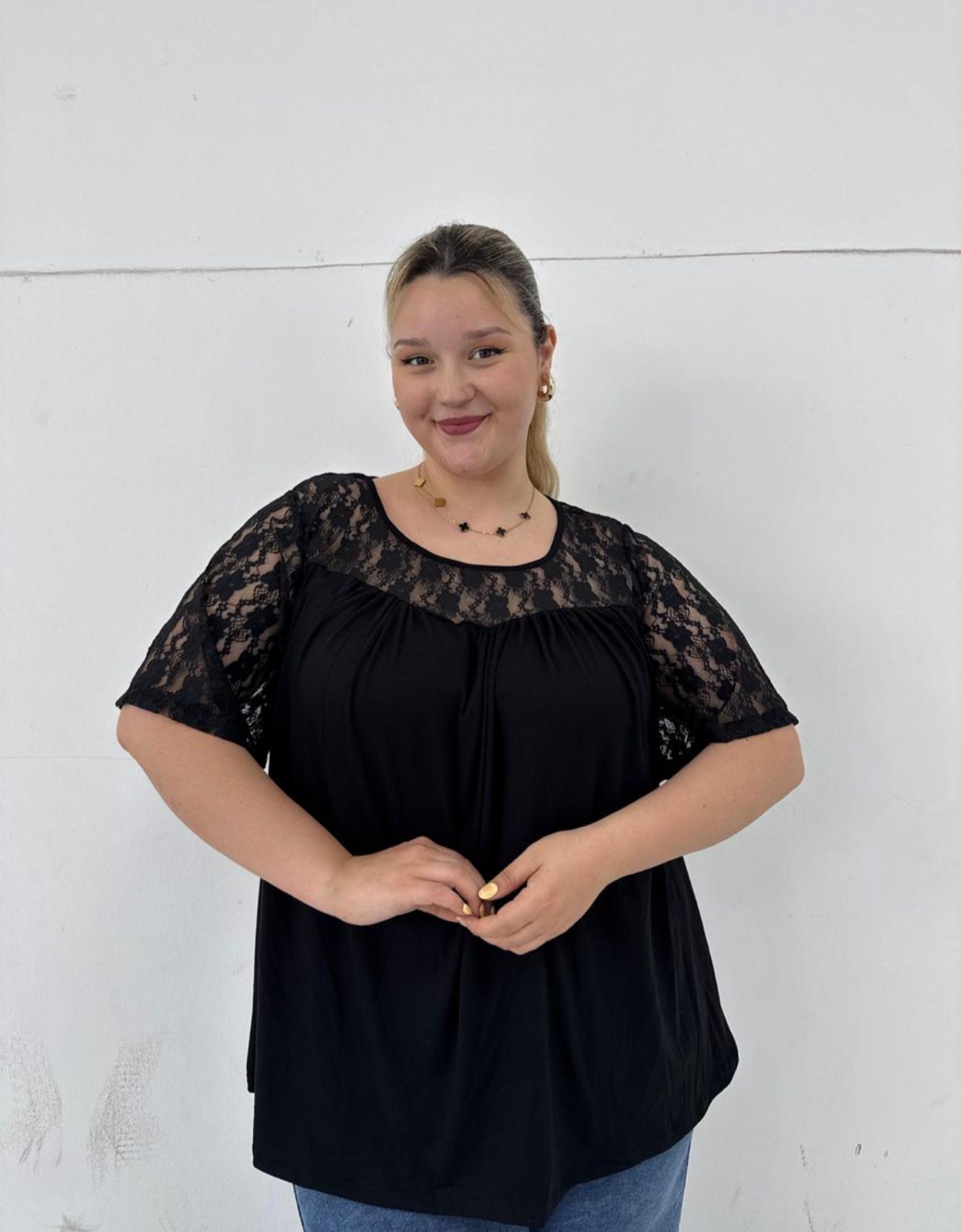 Mujer con top negro con mangas y escote de encaje, sosteniendo su camisa contra una pared blanca.