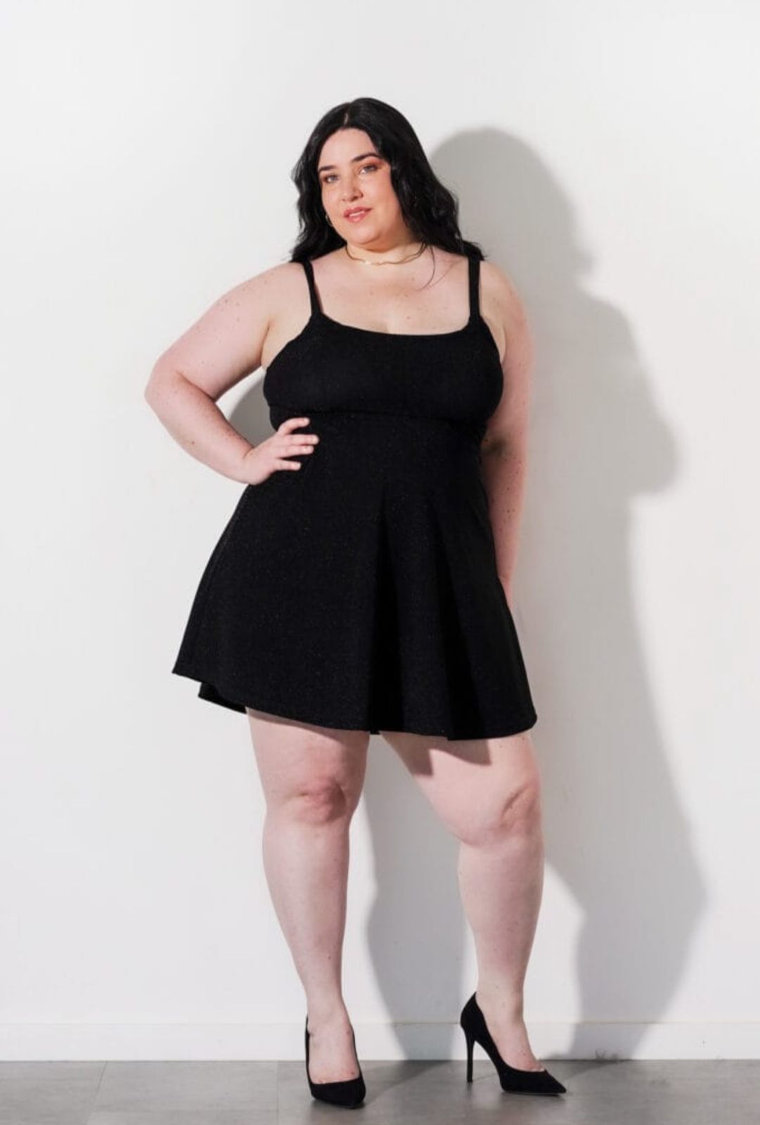 Mujer con un vestido negro brillante, de pie con las manos en las caderas, sonriendo, contra un fondo blanco.