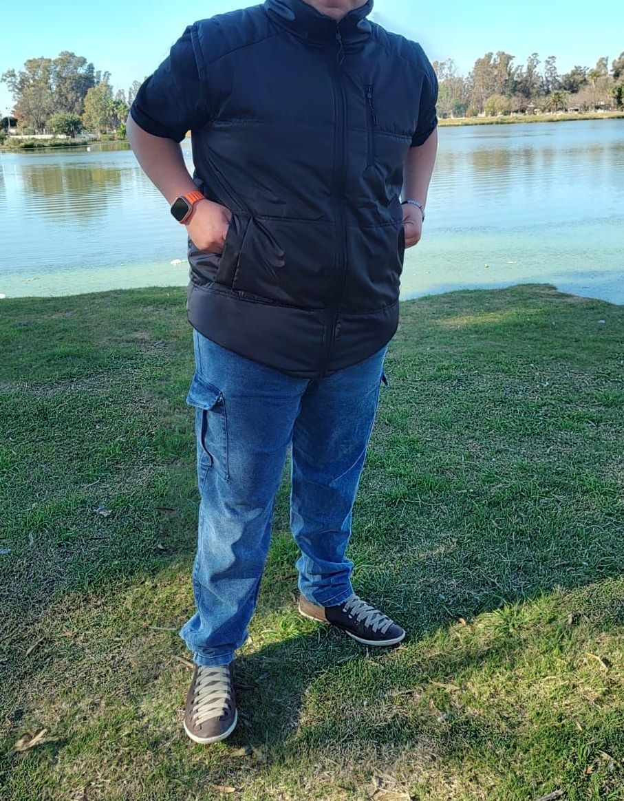 Un hombre vestido con un chaleco negro y pantalones vaqueros está parado junto a un lago en un día soleado.