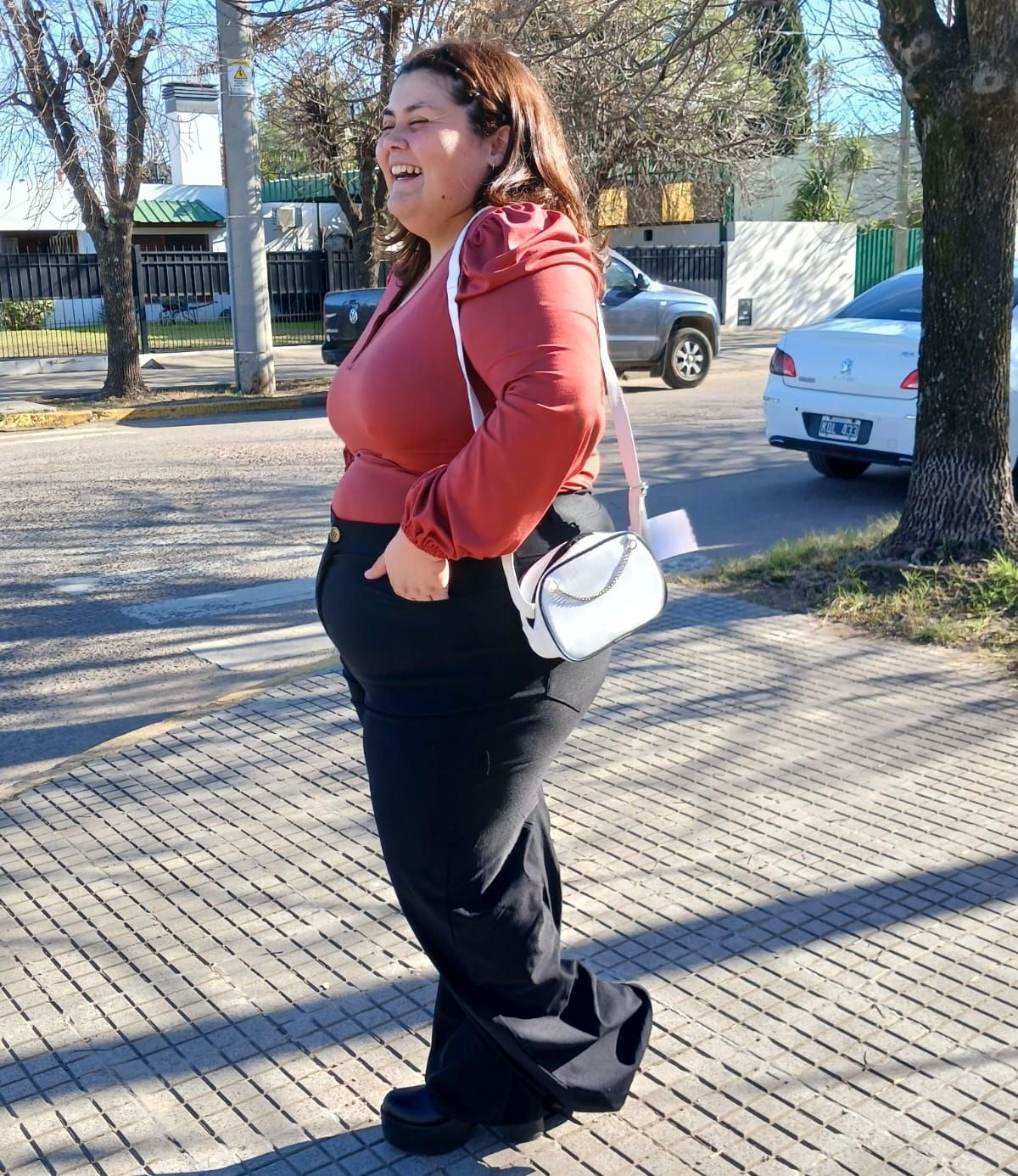 Una mujer con blusa color óxido y pantalones negros sonríe al cruzar la calle. Bolso blanco. Día soleado.