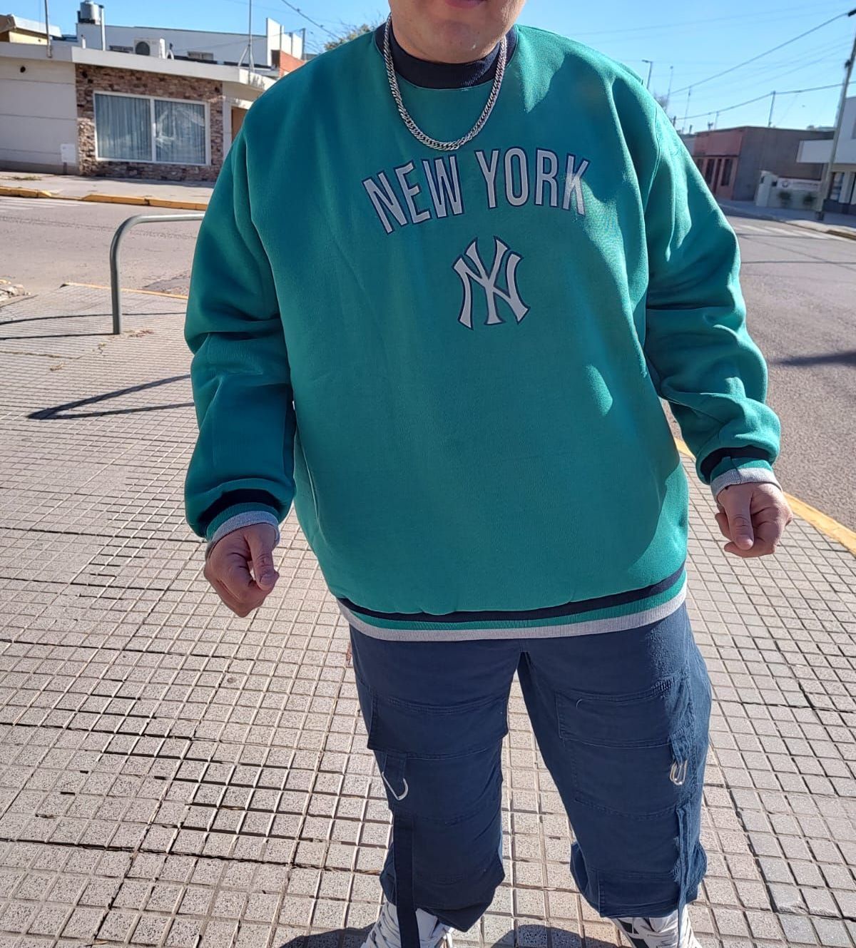 Hombre con sudadera verde azulado de Nueva York y pantalones deportivos azules de pie en una acera. Día soleado.