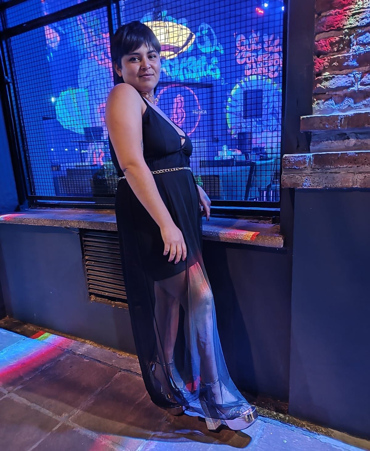 Mujer vestida de negro, sonriendo, en el interior cerca de una ventana con luces de colores.