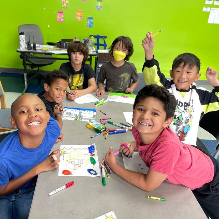 six boys drawing with markers while sitting around a table in front on a bright green wall