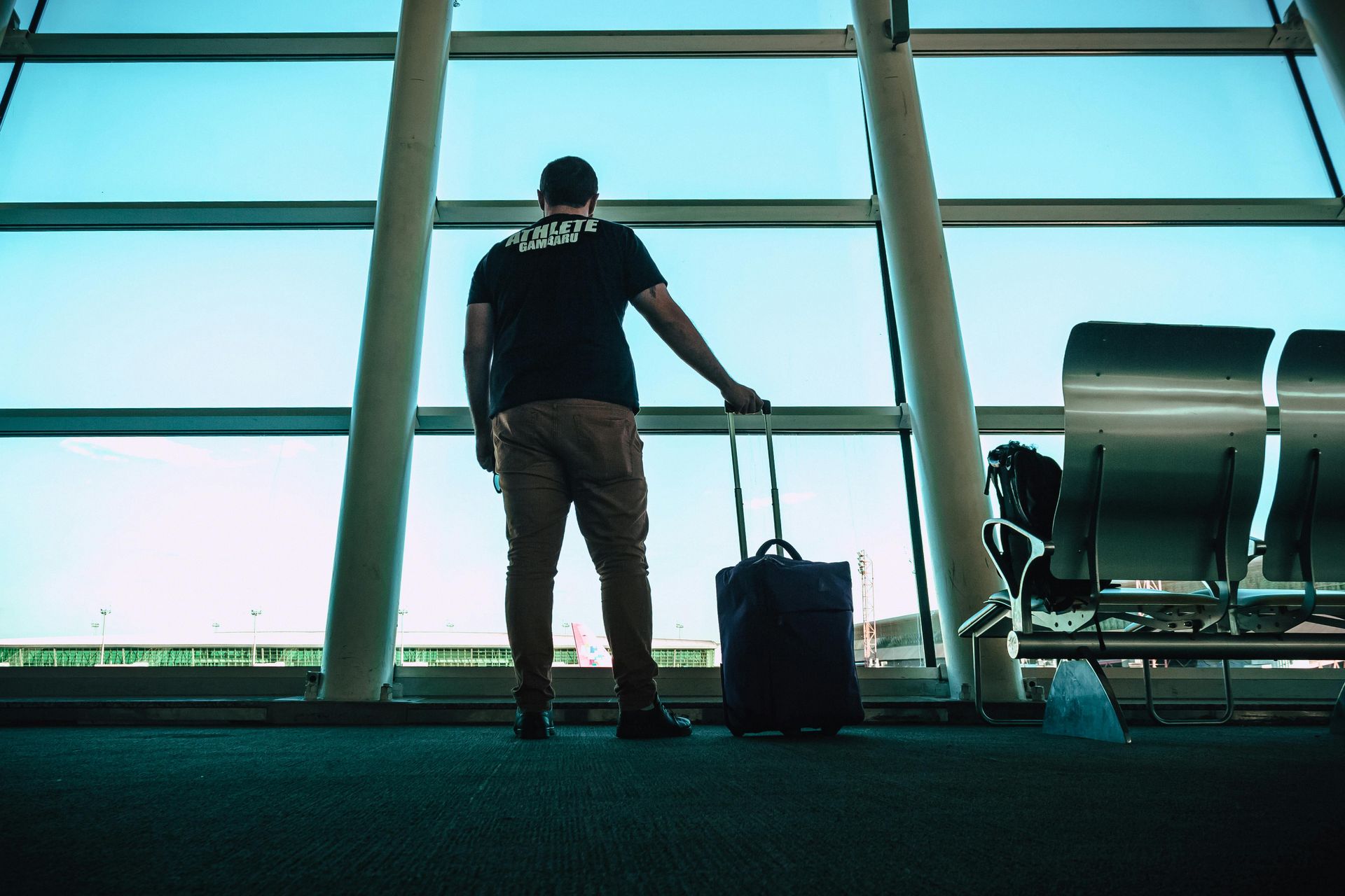 Man in black shirt stands at airport window, holding luggage.
