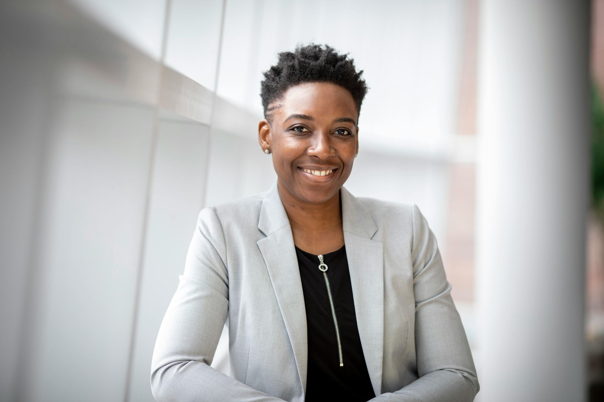 Smiling person with short, dark hair, in gray blazer, black shirt, against white backdrop.