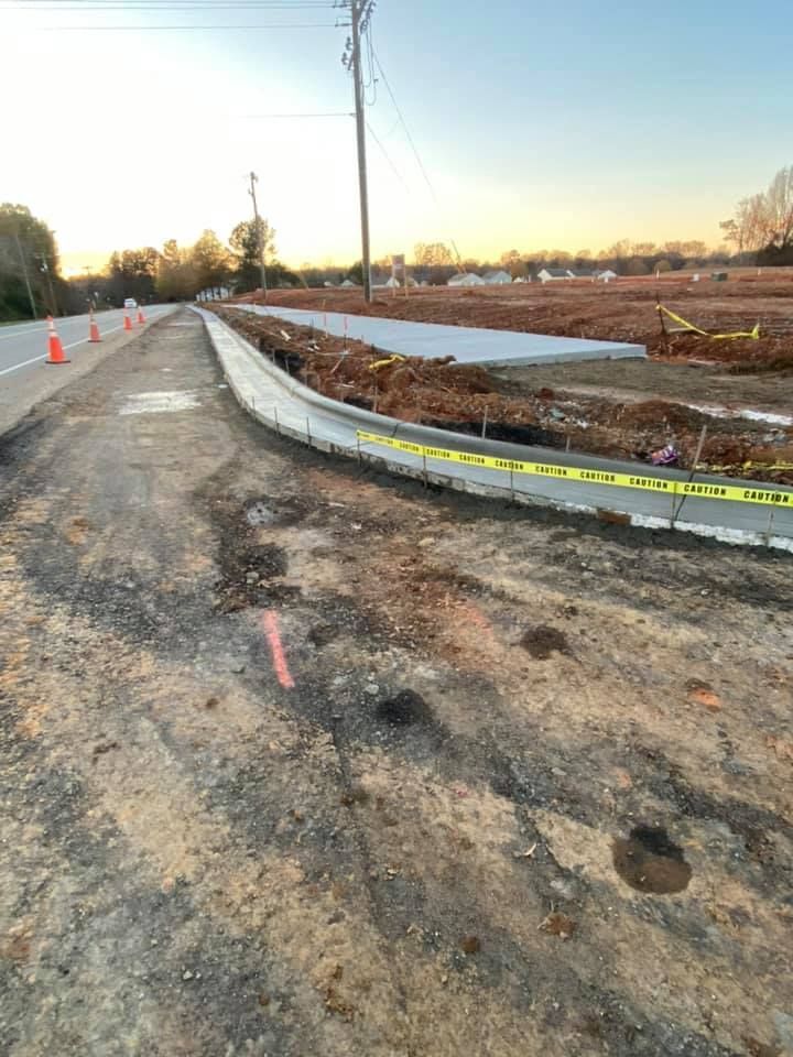 Road construction site. New curb, asphalt path, construction tape, and a field with trees and a utility pole.