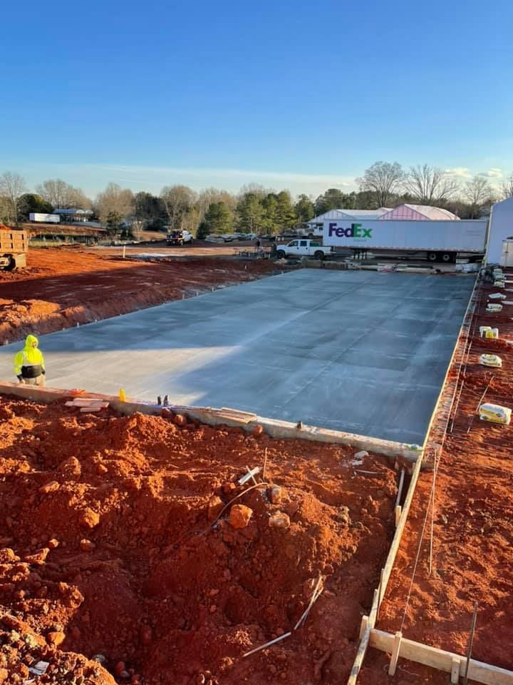 Construction site with freshly poured concrete slab. FedEx truck in background. Worker in yellow vest visible.