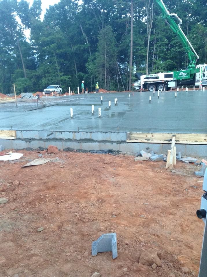 Construction site: concrete foundation being poured. Green concrete pump truck. Dirt foreground, trees in background.