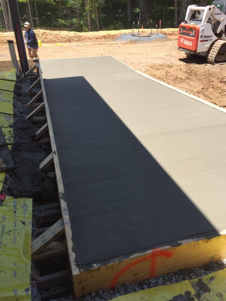 Freshly poured concrete walkway with wooden formwork at a construction site; person and Bobcat in the background.