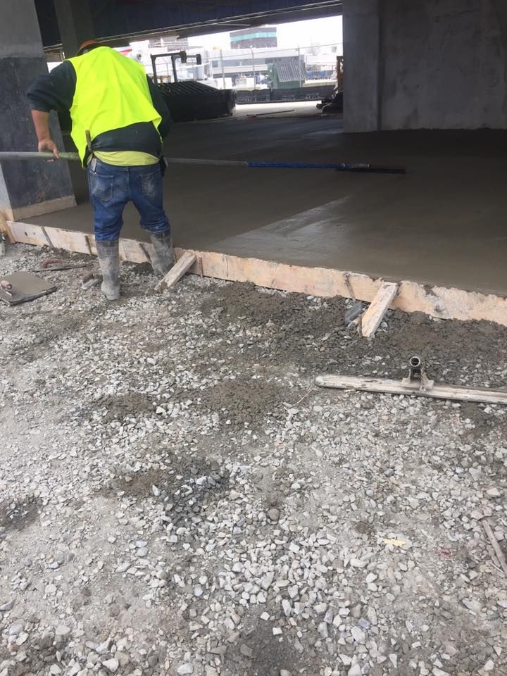 A worker in a yellow vest levels wet concrete with a long tool on a construction site.