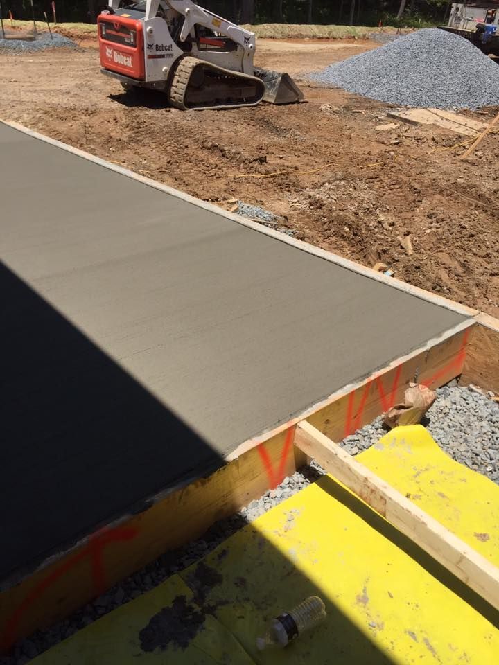 Freshly poured concrete walkway, framed with wood, with a Bobcat in the background at a construction site.