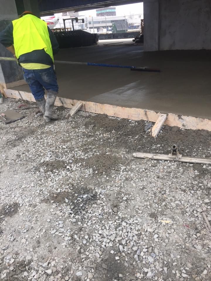 Construction worker smoothing wet concrete with a long tool. Yellow vest, concrete and gravel, under a bridge.