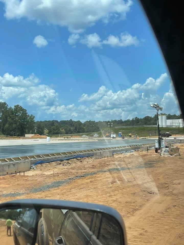 Construction site with concrete being poured on a sunny day. Blue sky, sandy ground, and workers visible.