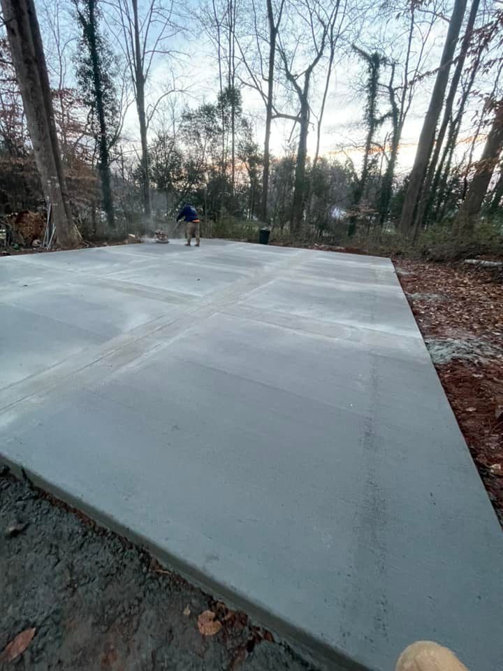 Newly poured concrete slab in a wooded area, person working in the background.