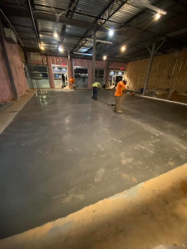 Workers smoothing fresh concrete floor in a dimly lit building with exposed beams.