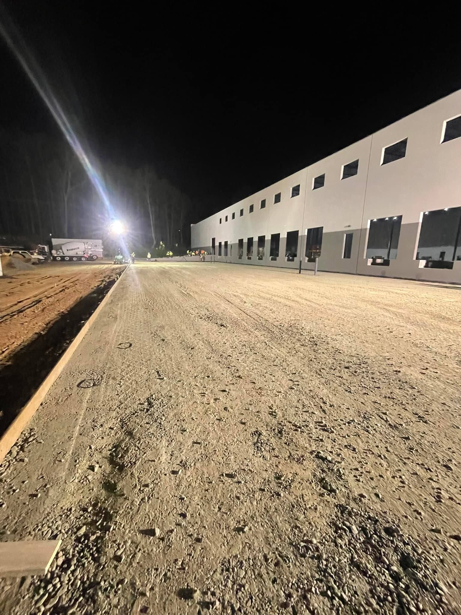 Construction site at night: gravel road leading to a long, white warehouse with multiple bay doors; lit by overhead lights.