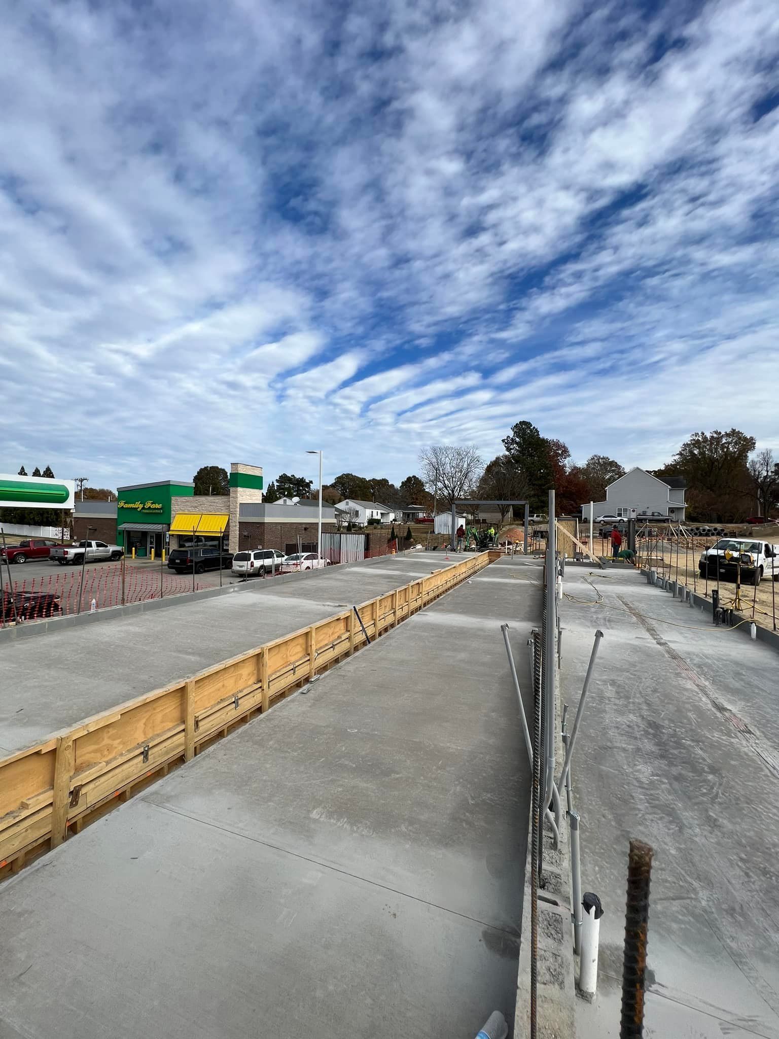 Construction site with concrete, wooden forms, and a cloudy blue sky. Buildings in the distance.
