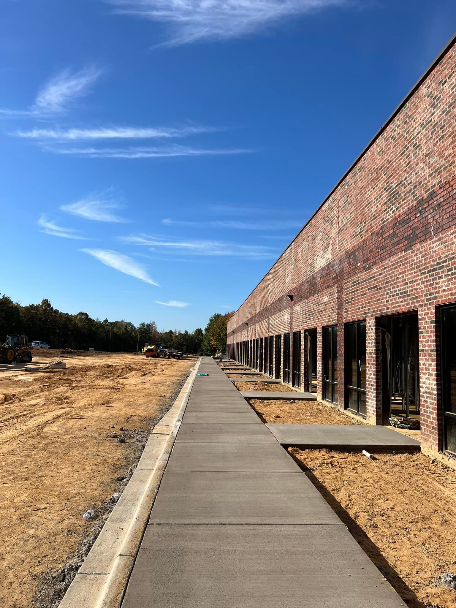 Long brick building under construction with a concrete walkway, bare earth, and blue sky.