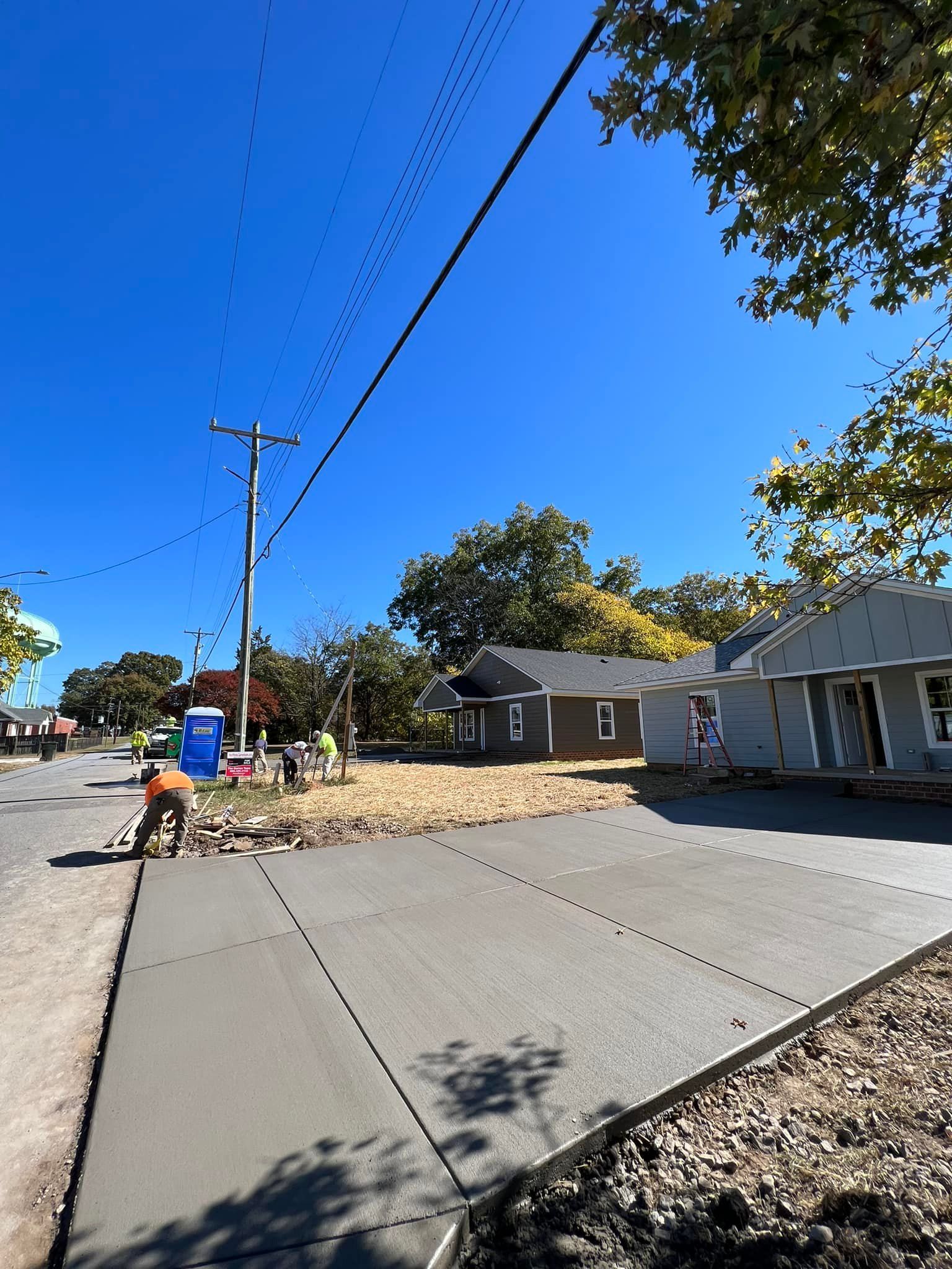 Construction site: Workers pouring concrete sidewalk under blue sky next to new houses and power lines.