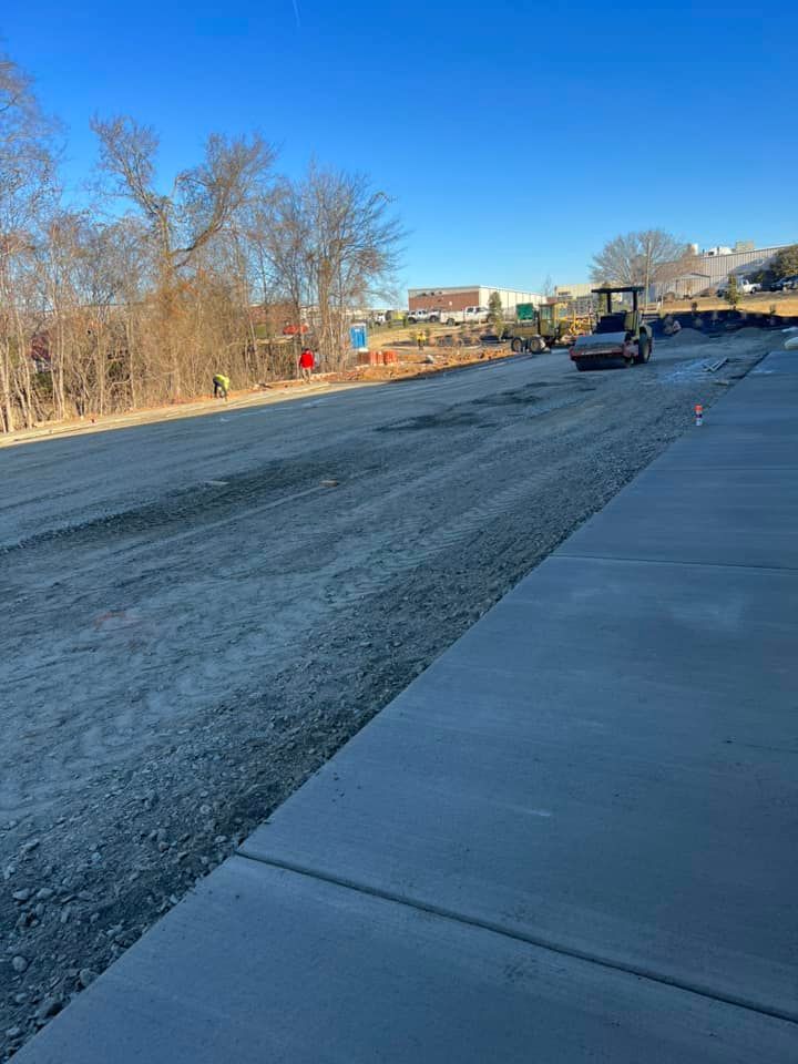 Construction site with a road under development. A roller compacts the gravel surface; sidewalk on the right.