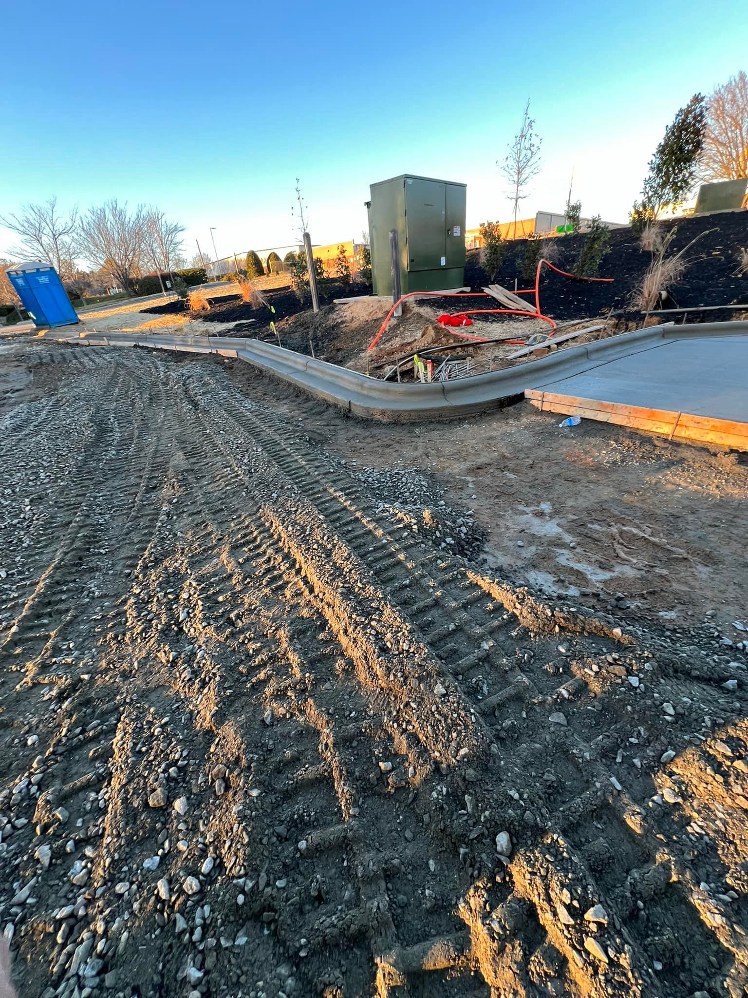 Construction site with gravel, dirt, and a newly poured concrete curb. An electrical box sits in the background.