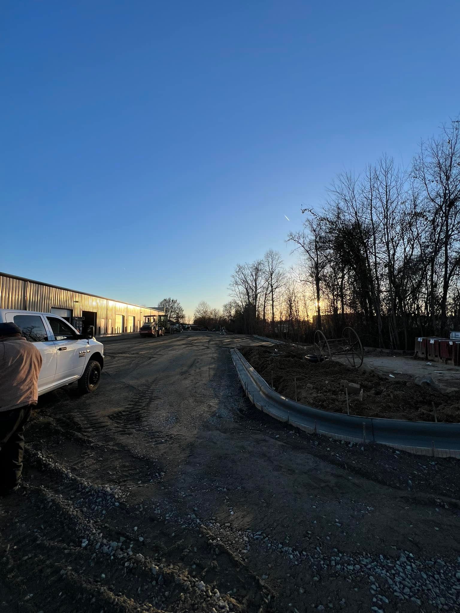 Construction site with a gravel road, parked truck, trees, and setting sun.
